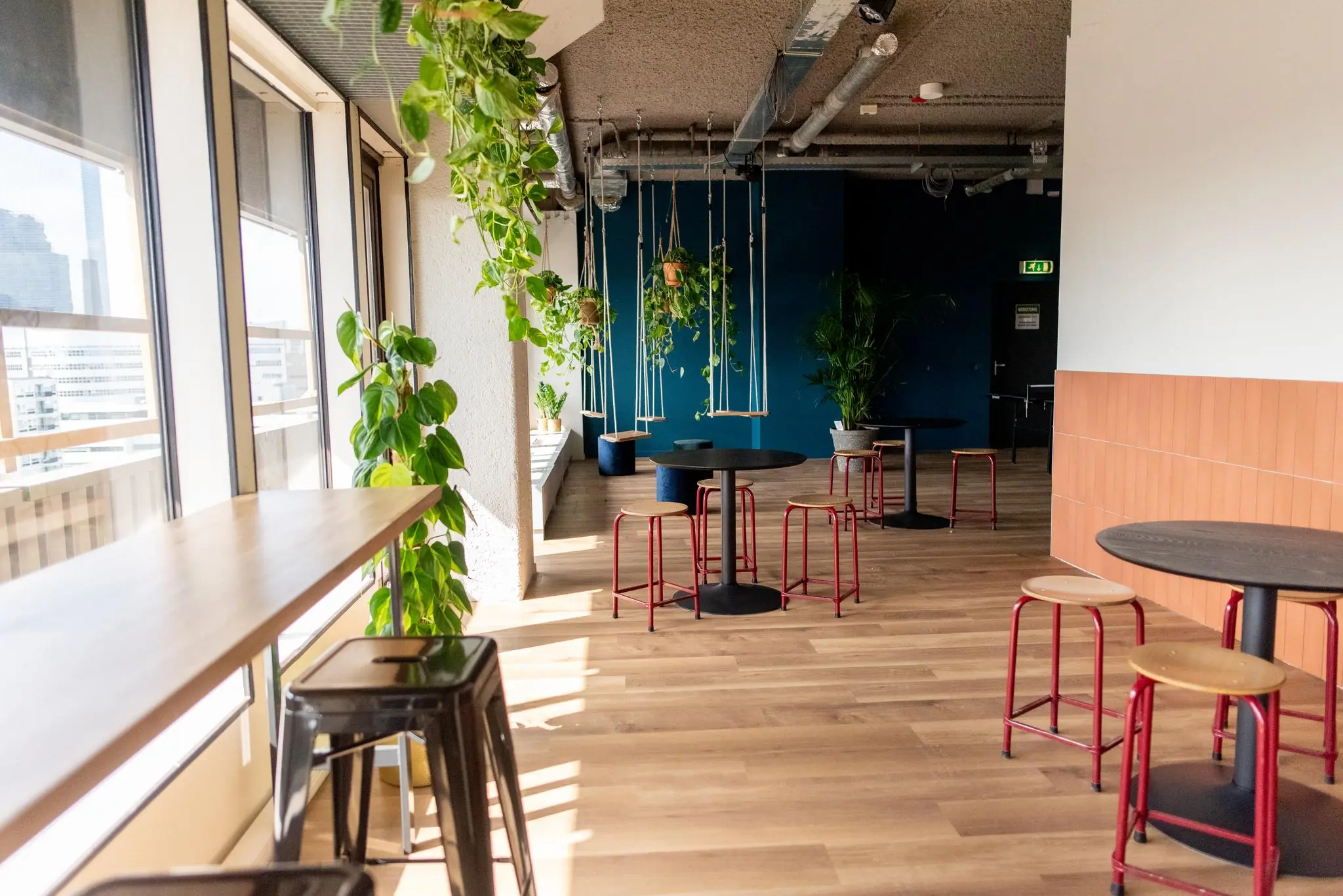 Moderne, lichte kantoorlounge aan de Aert van Nesstraat met zithoeken, hoge ramen en hangende planten.